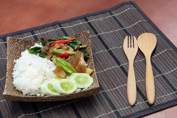 Thai food Stir-fried kale vegetable with crispy pork over rice. Placed on a wooden plate with natural pattern coconut. wooden spoons and forks on the small mat.