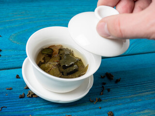 a bowl of brewed tea on a blue wooden background. A hand lifts the lid of the bowl. The opened leaves of large-leaved tea. Chinese tea. tea party