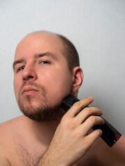 a man on a gray background shaves his beard with an electric razor. He looks at the camera, trimming his hair. Men's beauty and care at home.