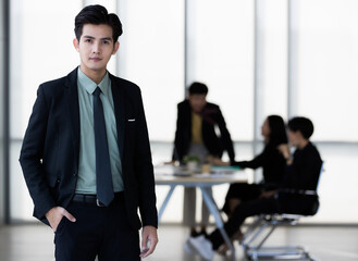 Young and handsome Asian businessman in body fit black suit standing pose in modern office with self-confidence and success gesture with other colleagues blur in background