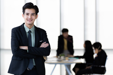 Young and handsome Asian businessman in body fit black suit standing pose in modern office with self-confidence and success gesture with other colleagues blur in background
