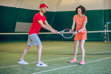 Young man in a red cap and with a racket having a workout with his female coach
