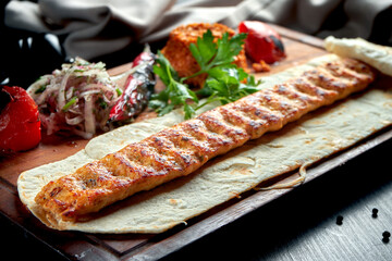 Grilled Turkish lamb lula kebab with grilled vegetables, onion and rice on a wooden board. Dark background.