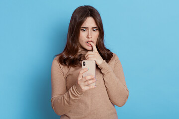 Gorgeous girl with dark hair holding phone in her hand, being surprised by something she saw on screen, has scared expression, biting her finger, wearing casually, isolated over blue background.