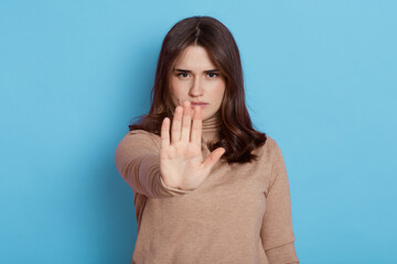 Beautiful girl showing stop sign with palms and looking directly at camera with serious expression, lady posing isolated over blue background, wearing beige turtleneck.