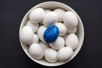 Blue egg lies on top of white ones in a bowl on black background. Easter or wealth concept. Minimalist