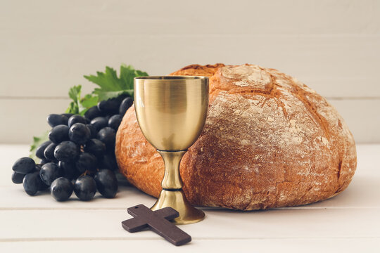 Chalice of wine with bread and cross on white wooden background