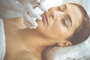 Beautician doing beauty procedure with syringe to face of young brunette woman in sunny clinic. Cosmetic medicine and surgery, beauty injections