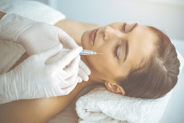 Obraz na płótnie Canvas Beautician doing beauty procedure with syringe to face of young brunette woman in sunny clinic. Cosmetic medicine and surgery, beauty injections