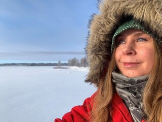 A happy woman in her 40's with a red winter coat enjoying a sunny day by the frozen sea or a lake.