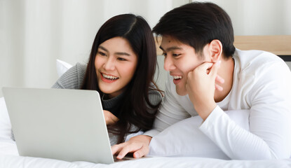 Young Asian black long hair woman and clear face man lovers laying on bed and using notebook computer to serf internet together with relax and happy