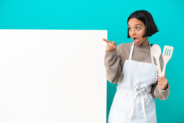 Young mixed race cook woman with a big placard isolated on blue background surprised and pointing up