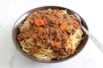 On a brown ceramic plate on a white marble table, typical spaghetti with minced beef, tomato sauce, carrot, and a fork. Selective focus, top view