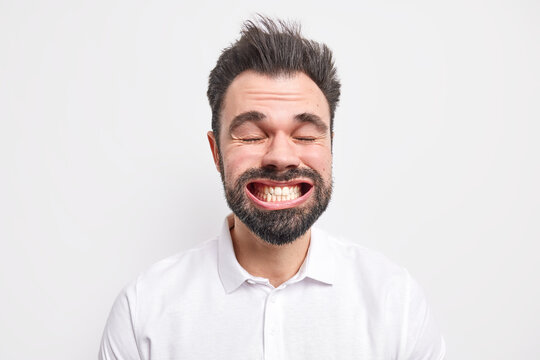 Headshot of funny bearded adult European man clenches teeth wears shirt closes eyes makes funny grimace dressed in shirt isolated over white background. Crazy mad unshaven guy with opened mouth