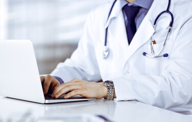 Unknown male doctor sitting and working with laptop in clinic at his working place, close-up. Young physician at work. Perfect medical service, medicine concept