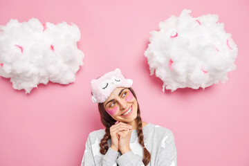 Portrait of good looking satisfied young woman looks with dreamy expression keeps hands under chin wears sleepmask and pajama isolated over pink background. People sleeping rest morning concept