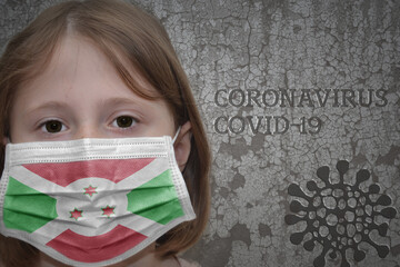 Little girl in medical mask with flag of burundi stands near the old vintage wall with text coronavirus, covid, and virus picture.