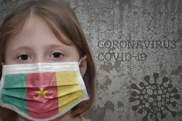 Little girl in medical mask with flag of cameroon stands near the old vintage wall with text coronavirus, covid, and virus picture.