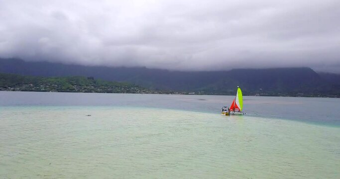 Aerial moving forward along a sandbar with a catamaran, small boat, cloudy skies, and rugged mountains in the background - Oahu, Hawaii