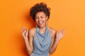 Overjoyed positive young Afro American woman has trendy hairstyle dressed in polka dot blouse smiles broadly raises palms has fun on party closes eyes poses against vivid orange studio background