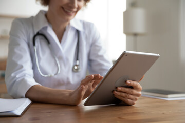 Crop close up female doctor hold tablet gadget consult patient online. Woman GP sit at desk in hospital use pad device browse surf wireless internet, text or message. Medicine and technology concept.