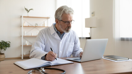 Serious mature Caucasian male doctor in white medical uniform sit at desk in hospital work on computer write in journal. Elderly man GP or therapist consult client online on laptop note in notebook. - obrazy, fototapety, plakaty