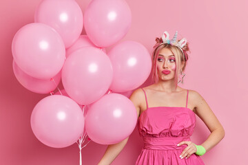 Obraz na płótnie Canvas Pensive woman with hairstyle dressed in festive dress holds bunch of balloons thinks about decorations for party awaits for starting event isolated over pink background. Special occasion concept