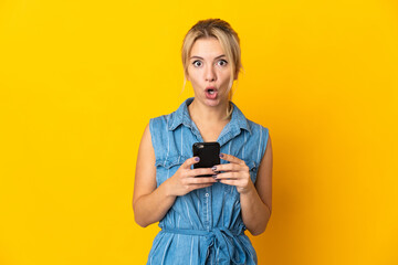 Fototapeta premium Young Russian woman isolated on yellow background looking at the camera while using the mobile with surprised expression