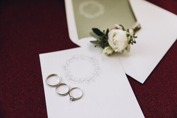 Wedding details. The rings of the bride and groom lie on a white invitation card and an envelope, next to it lies a boutonniere of white flower and greenery
