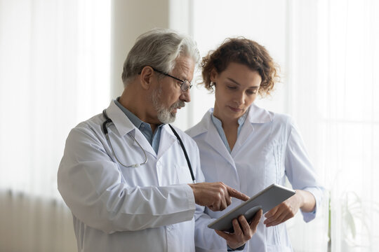 Focused Mature Male Doctor And Female Nurse Look At Tablet Screen Discuss Anamnesis Together. Concentrated Diverse Medical Professionals Use Pad Device, Engaged In Team Thinking In Hospital.