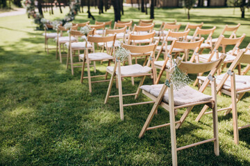 Wedding ceremony. On a green meadow in the forest there is an arch of flowers and greenery, next to there are wooden chairs for guests