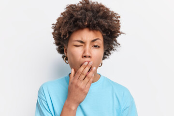 Fototapeta na wymiar Headshot of young Afro American sleepy woman yawns has exhausted expression being tired of everything fed up of working wears casual blue t shirt isolated over white background. Lack of energy
