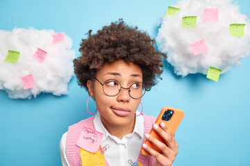 Close up shot of thoughtful curly haired busy young woman holds mobile phone concentrated aside checks email box works in office makes stickers with information to remind. Adhesive notes around