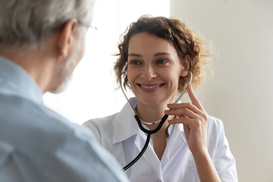 Close up of smiling young Caucasian female doctor use stethoscope examine mature patient heart rate in clinic. Happy caring woman nurse hold phonendoscope listen to elderly man heartbeat in hospital.