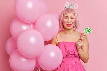 Crying mournful woman has pink wig feels unhappy on party holds sweet candy on stick and bunch of inflated balloons wears dress isolated over rosy background. People holidays bad mood concept