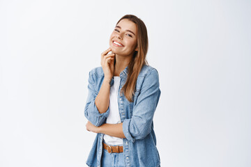 Portrait of beautiful young woman with perfect skin, natural make up, touching face and smiling at camera, standing half-turned against white background