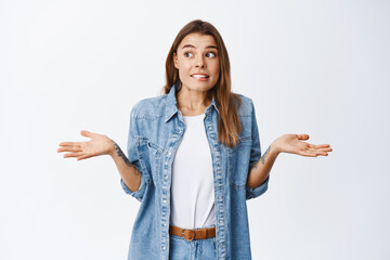 Oops sorry. Clueless young woman shrugging and looking away unsure, dont know and have no idea, nothing to tell, standing against white background - obrazy, fototapety, plakaty