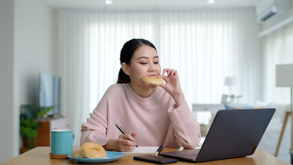 Young attractive beautiful asian female hungry eat doughnut take away snack food with full mouth look at computer notebook at home in busy work from home multitask unhealthy meal lifestyle concept.