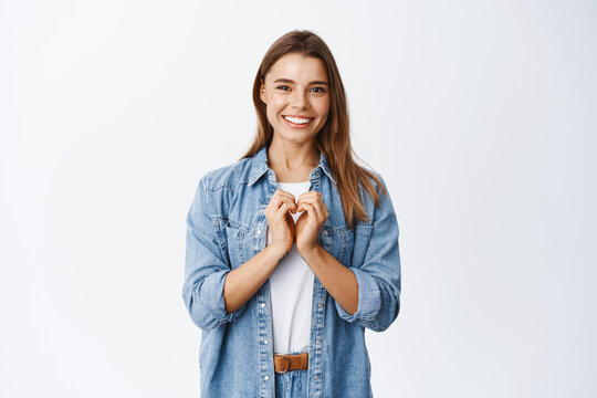 Beautiful romantic girl showing heart gesture, I love you sign, smiling happy at camera, standing against white background