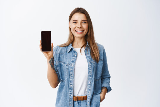 Portrait of attractive young woman with white smile, showing empty smartphone screen, demonstrate an app or online shopping store, studio background