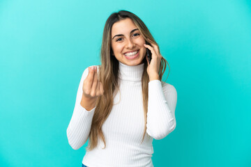 Fototapeta premium Young caucasian woman using mobile phone isolated on blue background making money gesture
