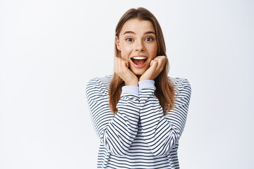 Fototapeta premium Portrait of fascinted and amazed blond girl looking at camera fascinated, checking out something cool, seeing interesting thing, standing over white background