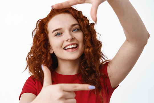 Close up portrait of beautiful ginger girl with curly hair and natural light make up, making hand frames photo gesture and smiling, picturing something, white background