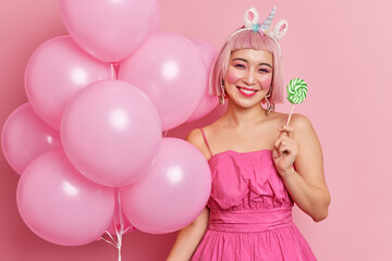 Obraz na płótnie Canvas Positive Asian girl smiles gently has bright makeup wears dress holds delicious sweet candy bunch of inflated balloons enjoys holiday celebrates birthday being on party isolated over pink background