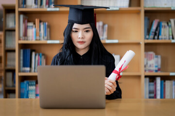 A young Asian female university graduate expressing joy and excitement to celebrate her achievement of degree graduation in front of a laptop making a remote video call to her parents at home