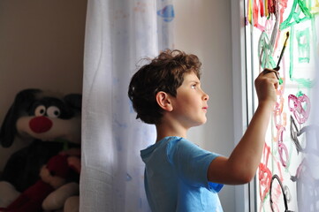 A little serious boy holds a brush in his hands and draws pictures on the window in his room. The child draws a rainbow, a house and an abstraction. Stay at home. Plush bunny spying on a boy.