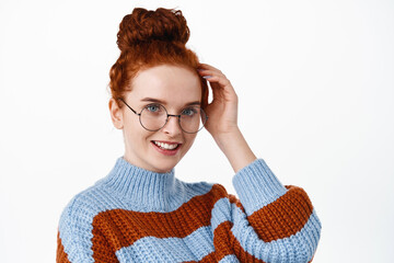 Close up of beautiful and romantic woman with ginger hair combed in hairbun, wearing glasses, touching hair and smiling flirty, blushing cute, standing against white background