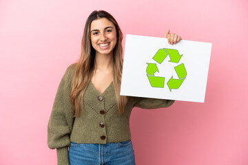 Young caucasian woman isolated on pink background holding a placard with recycle icon with happy expression