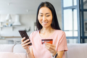 Cheerful young Asian woman making an order online, holds a smartphone and a debit cad, pays for the purchase by the phone, a multiracial girl is using mobile phone for online shopping