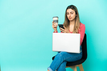 Young caucasian woman sitting on a chair with her pc isolated on blue background holding coffee to take away and a mobile